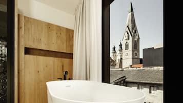 Un baño moderno con una bañera independiente, paneles de madera y vistas a una torre de iglesia a través de un ventanal grande.