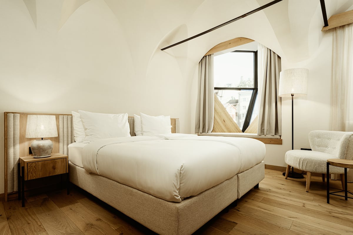 A modern bedroom with a large bed, a nightstand with a lamp, a chair, and a window with curtains.