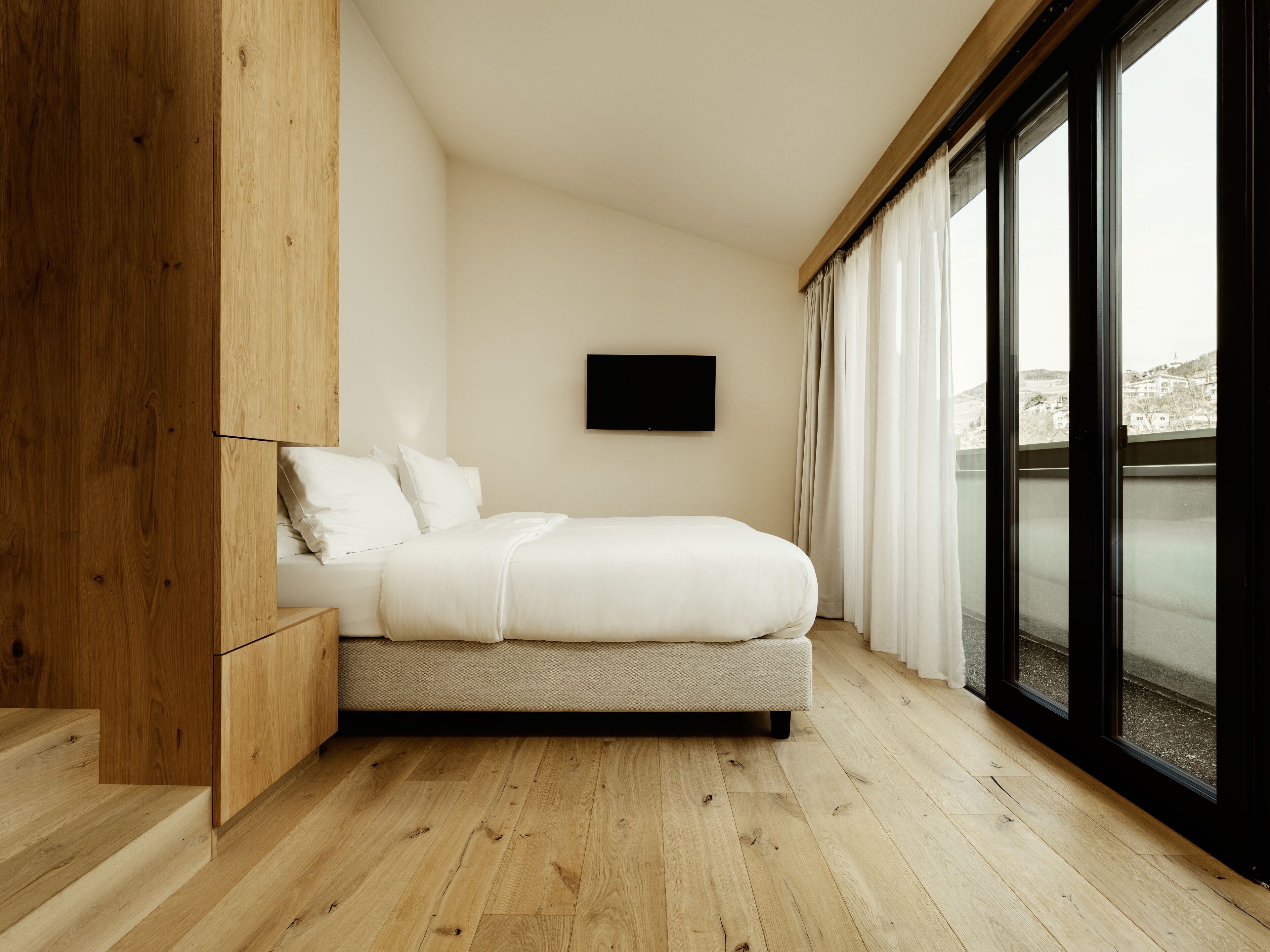 Un dormitorio moderno con suelo de madera, una cama con ropa de cama blanca, un televisor montado y ventanas grandes con cortinas.