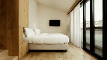 Un dormitorio moderno con suelo de madera, una cama con ropa de cama blanca, un televisor montado y ventanas grandes con cortinas.