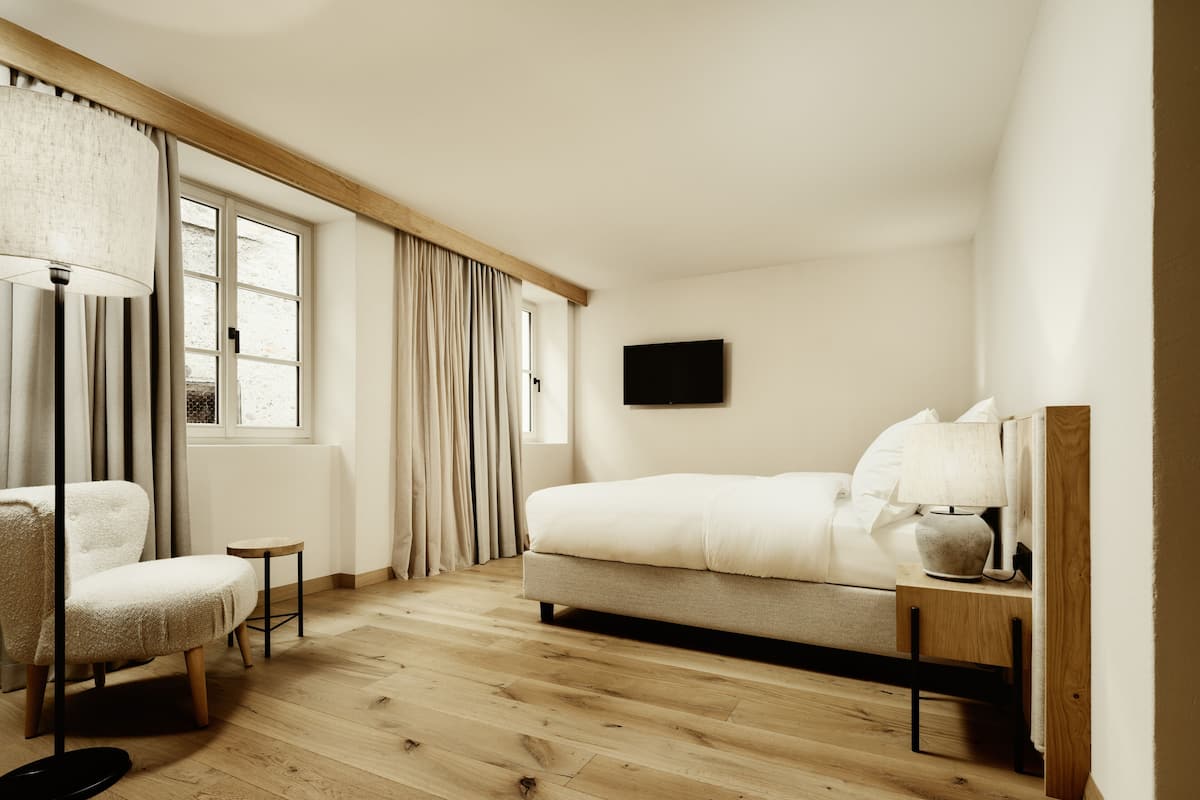 A bedroom with a bed, a chair, a bedside table, a lamp, and a TV on the wall.