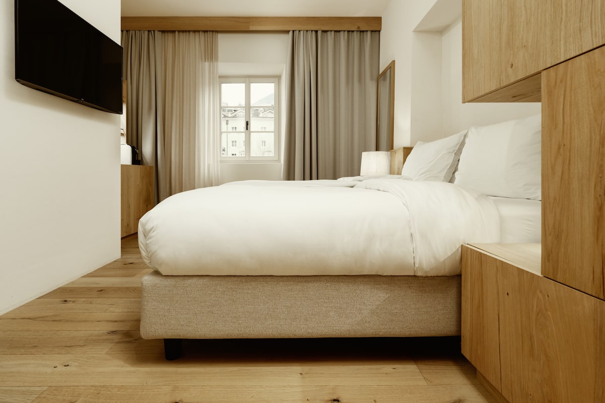 A modern hotel room with a large bed, wooden headboard, a flat-screen TV mounted on the wall, and a window with curtains.