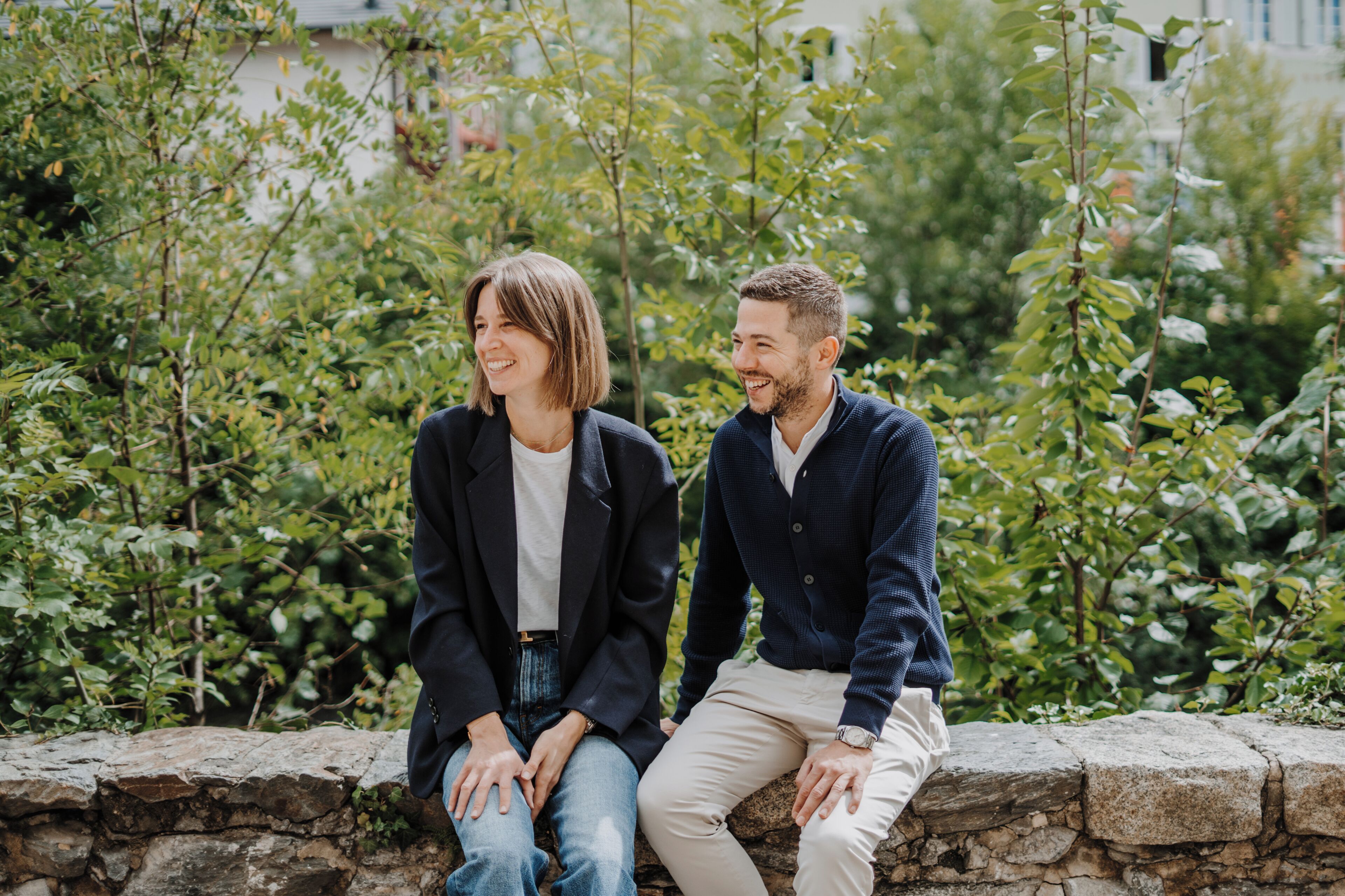 Two people sitting on a stone wall in a garden with greenery in the background.