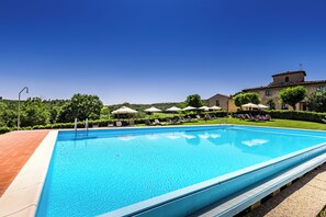 Een rechthoekig zwembad met helderblauw water, omgeven door een betegeld zonnedek. Er staan loungebedden en parasols langs het zwembad, en op de achtergrond staat een gebouw met een rood dak.