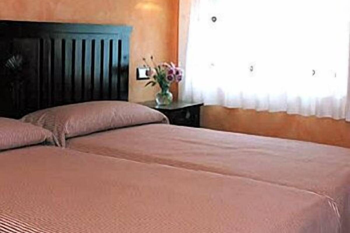 A bedroom with two beds, a nightstand with a vase of flowers, a wall-mounted light, and a window with a curtain.