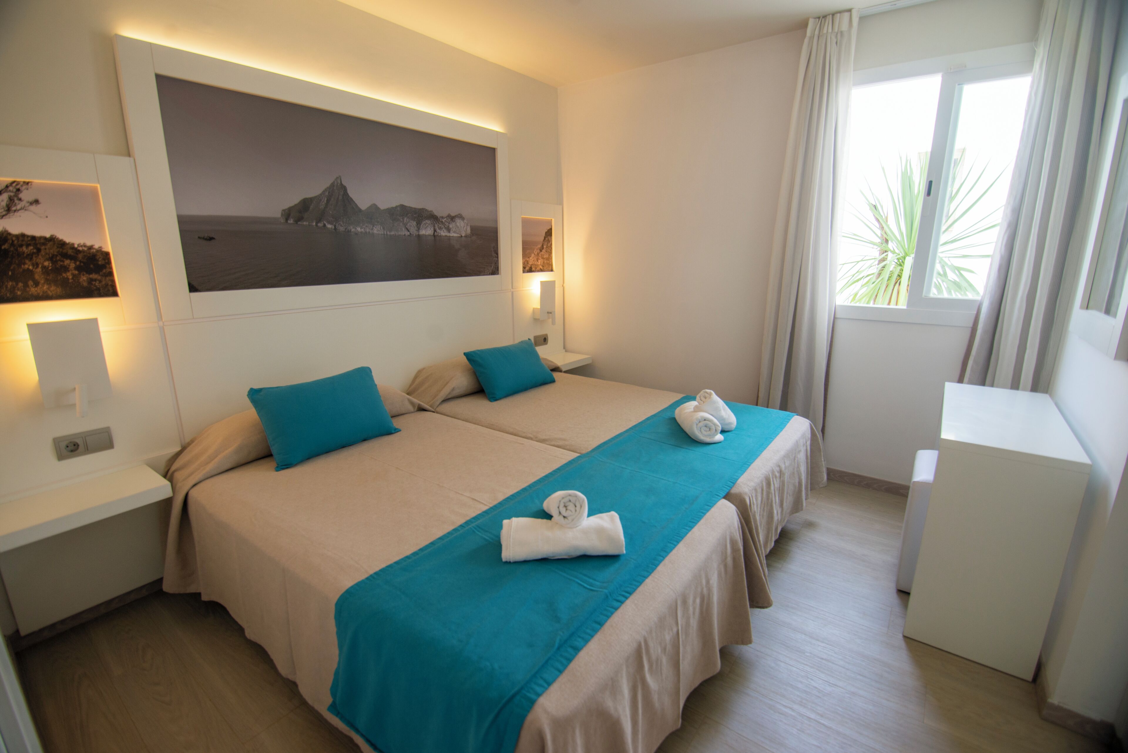 A hotel room with two beds, a large framed picture on the wall, a desk, and a window with curtains.