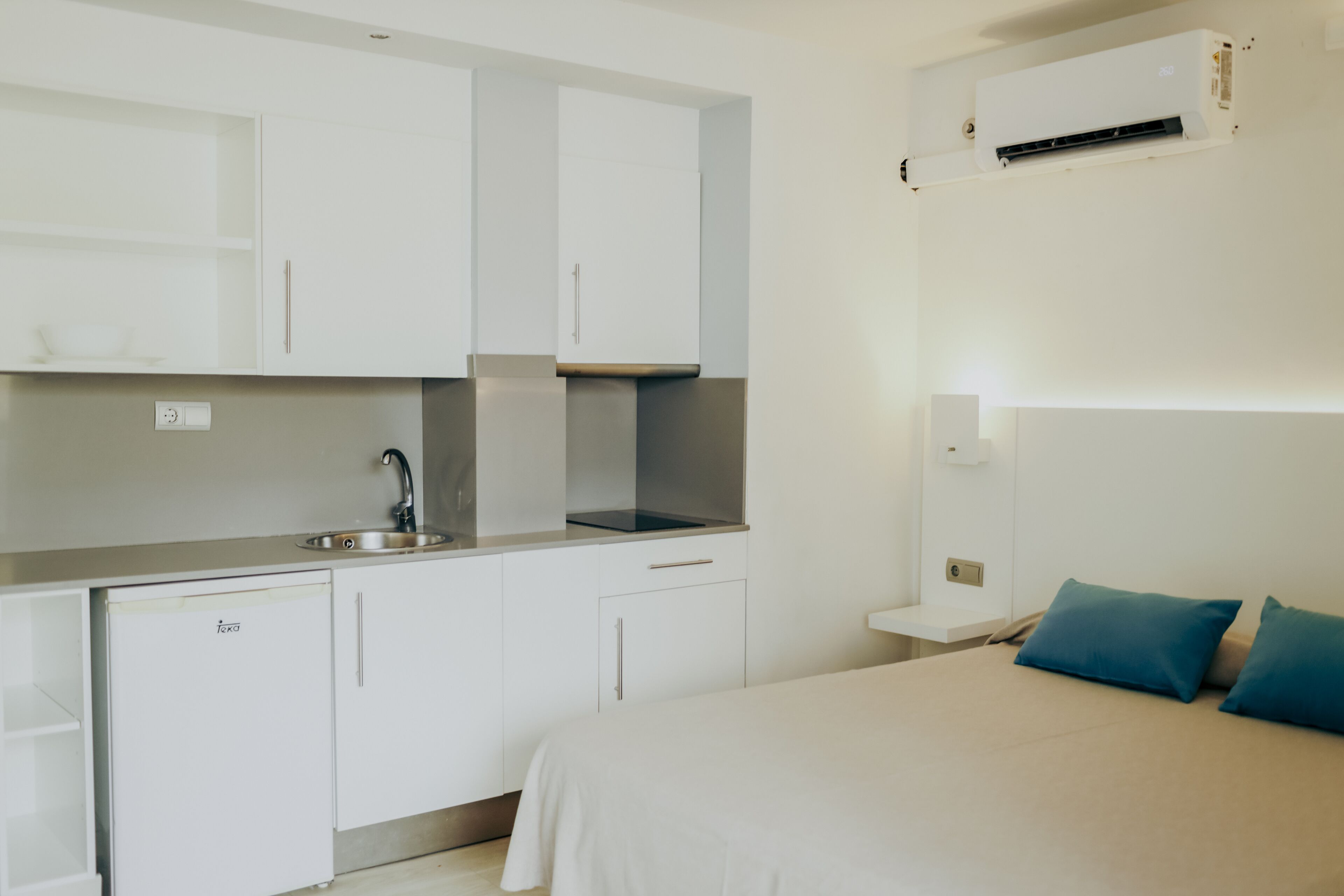 Une chambre d’hôtel compacte, dotée d’une kitchenette, d’un lit avec des oreillers de couleur turquoise et d’un climatiseur fixé au mur.