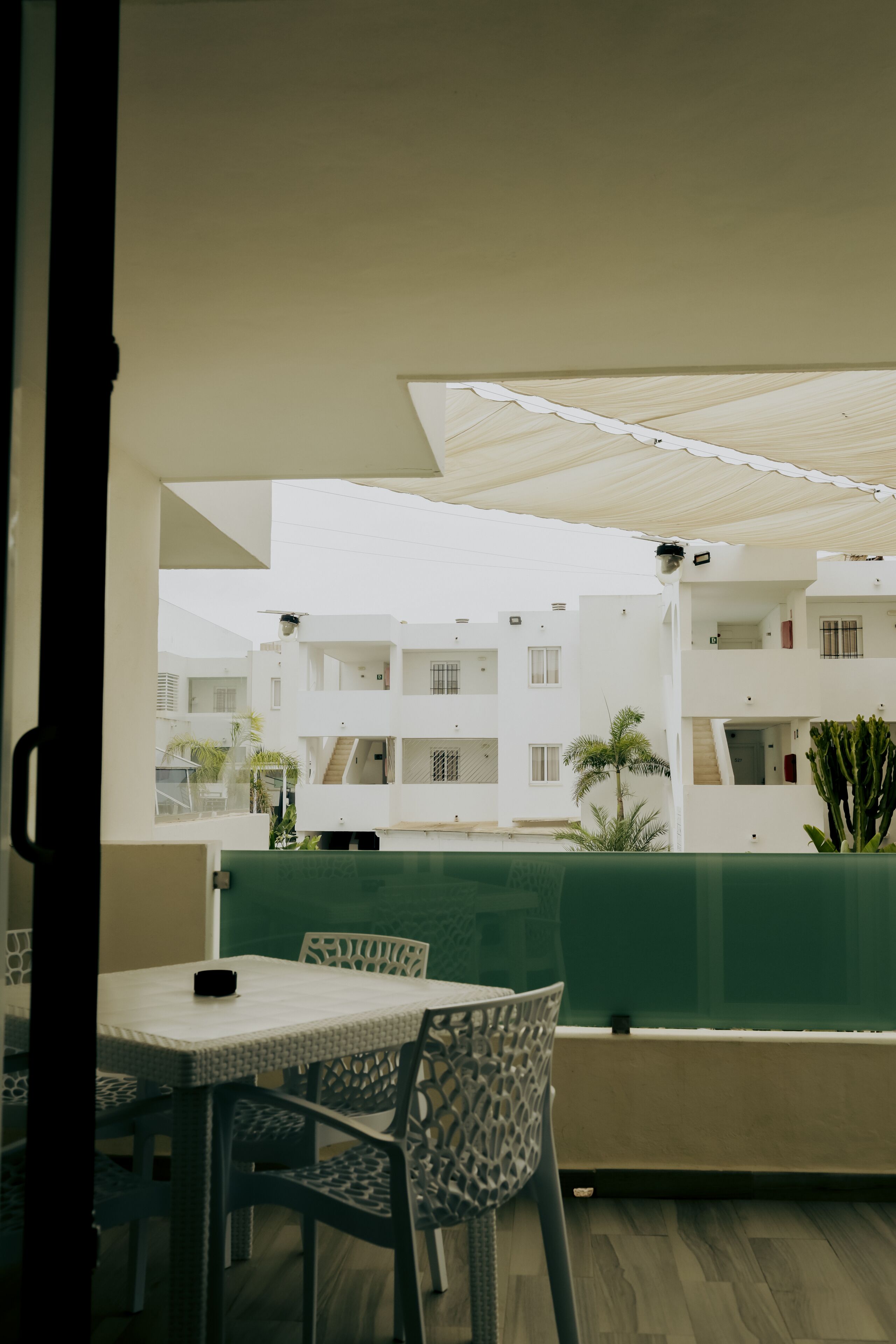Uma área de estar ao ar livre com mesa e cadeiras, um banco acolchoado verde e vista para um prédio branco com várias varandas.