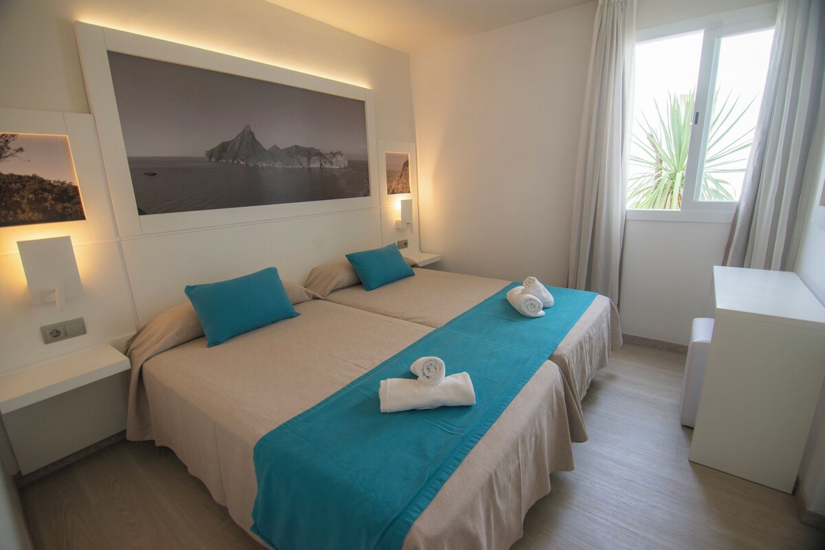 Ein Hotelzimmer mit zwei Betten, einem großen Wandbild, einem Nachttisch mit Lampe und einem Fenster mit Vorhängen.