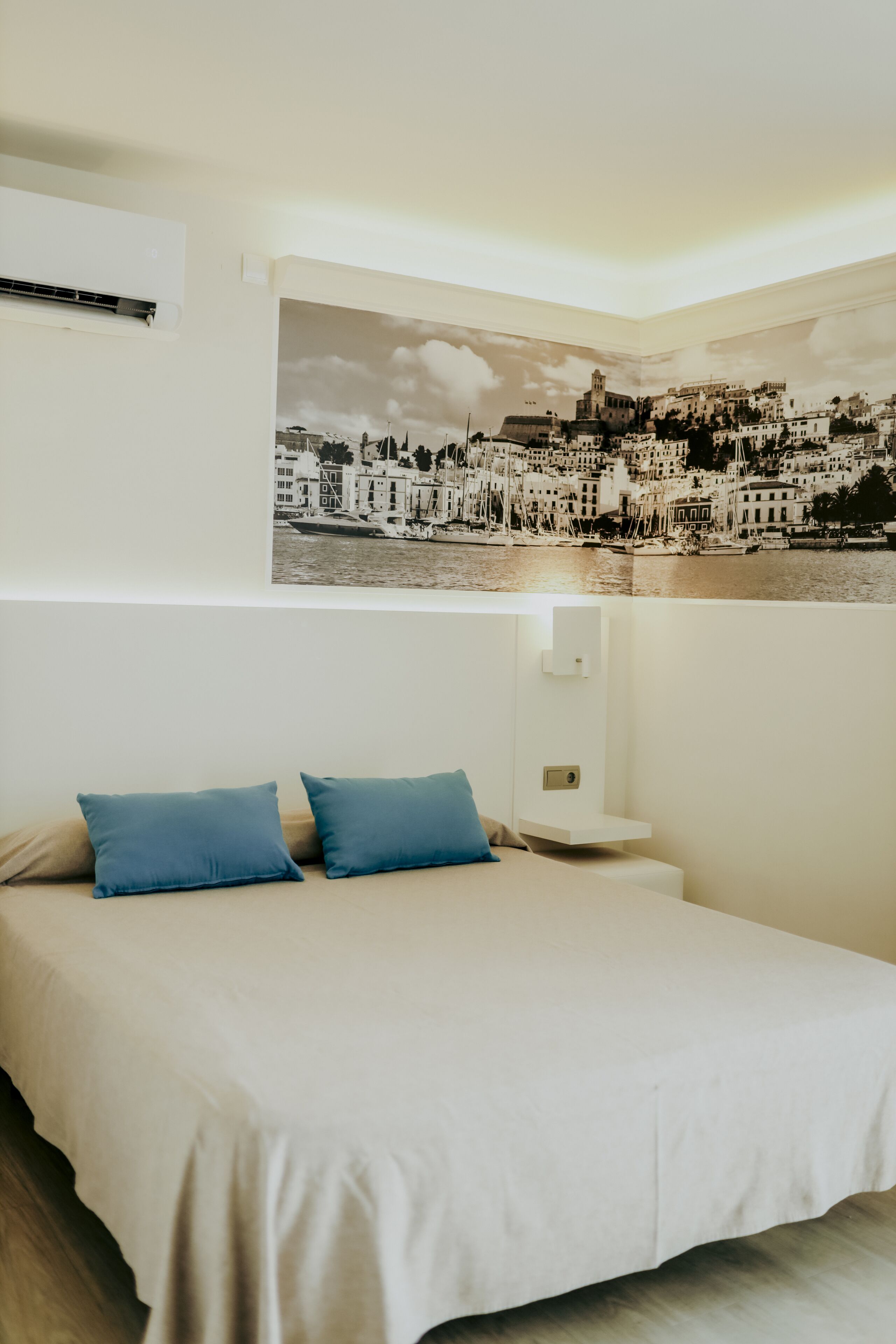 Un dormitorio con una cama, dos almohadas azules, una mesita de noche montada en la pared y un cartel grande con una vista urbana.