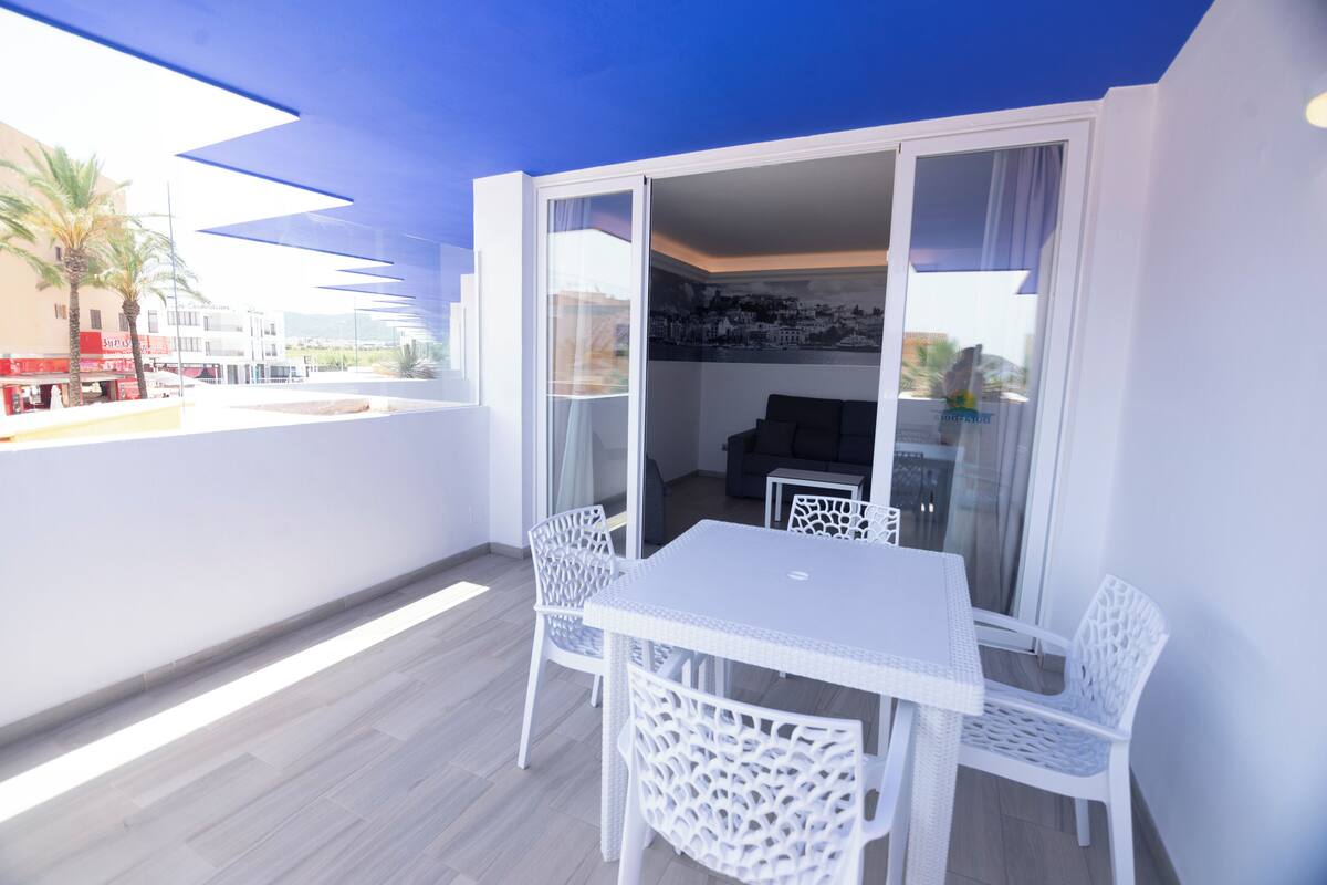 Un balcón con una mesa y sillas blancas, vistas de la ciudad y una puerta que da a un espacio interior.