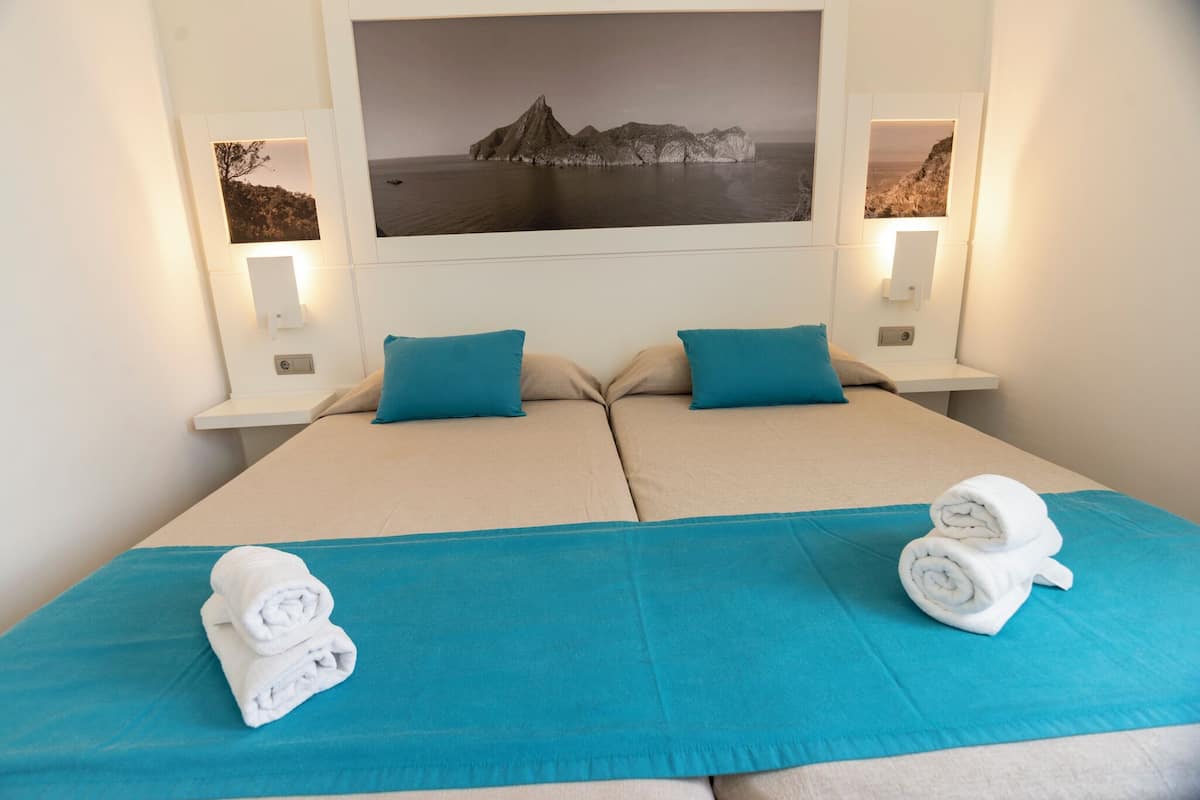 Una cama doble con dos almohadas, una colcha azul y dos toallas enrolladas sobre la cama.