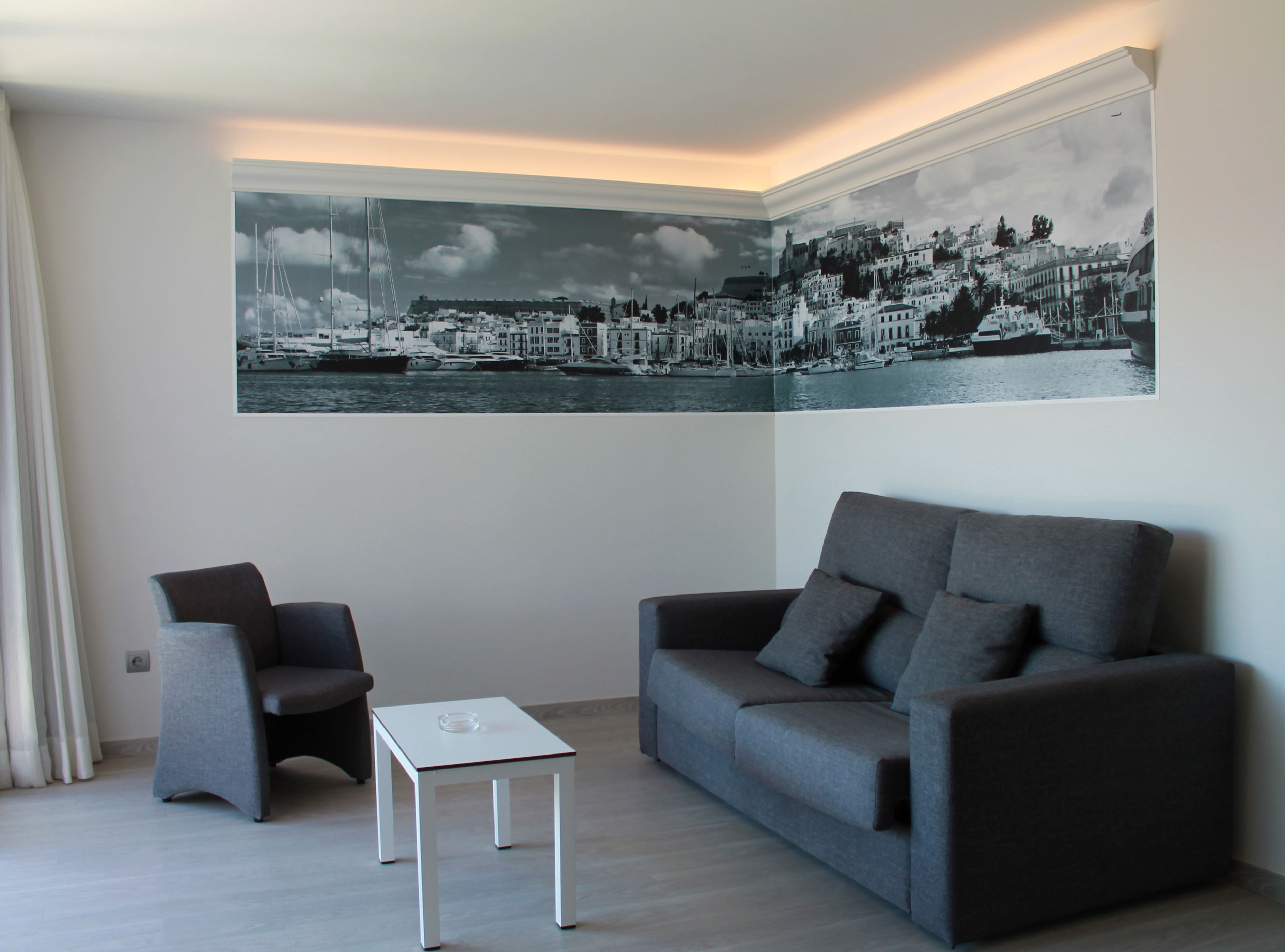 Ett modernt vardagsrum med en stor väggmålning som föreställer en svartvit stadsbild, en grå soffa, en grå fåtölj och ett litet vitt bord.