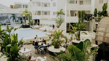 Zona de piscina en la azotea con sillones de descanso y mesas, rodeada de plantas en macetas y un edificio moderno al fondo.