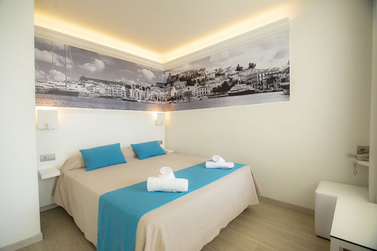 Habitación de hotel con cama, dos almohadas, colcha azul, un gran mural de paisaje urbano en la pared y una mesita de noche pequeña.
