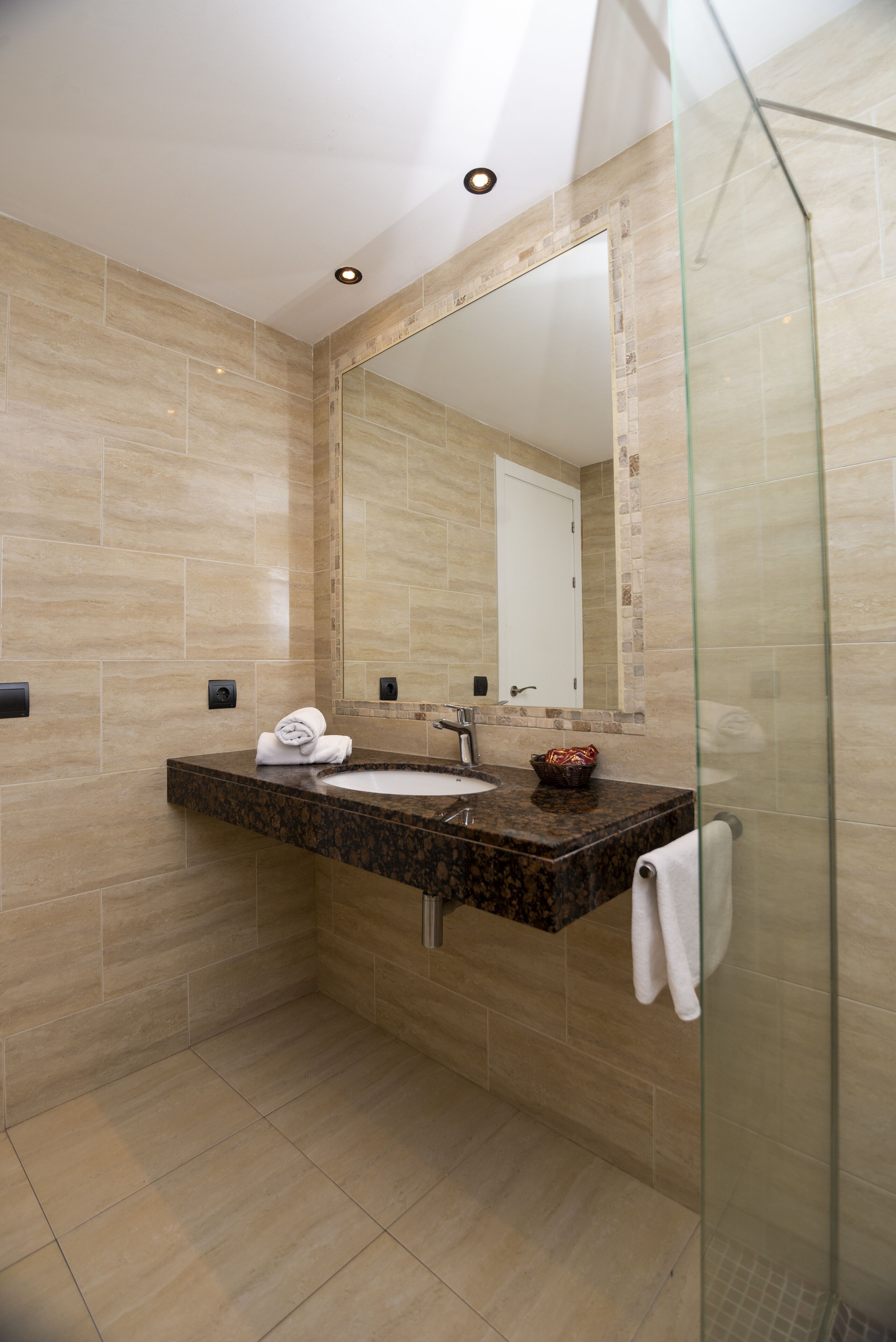 Une salle de bain moderne avec un lavabo surmonté d’un plan de travail en granit, une douche en verre et des murs recouverts de carreaux beiges.