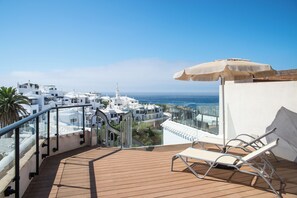 Une terrasse en bois, dotée de chaises longues et d’un parasol, qui offre une vue sur une ville côtière aux bâtiments blancs et sous un ciel bleu azur.