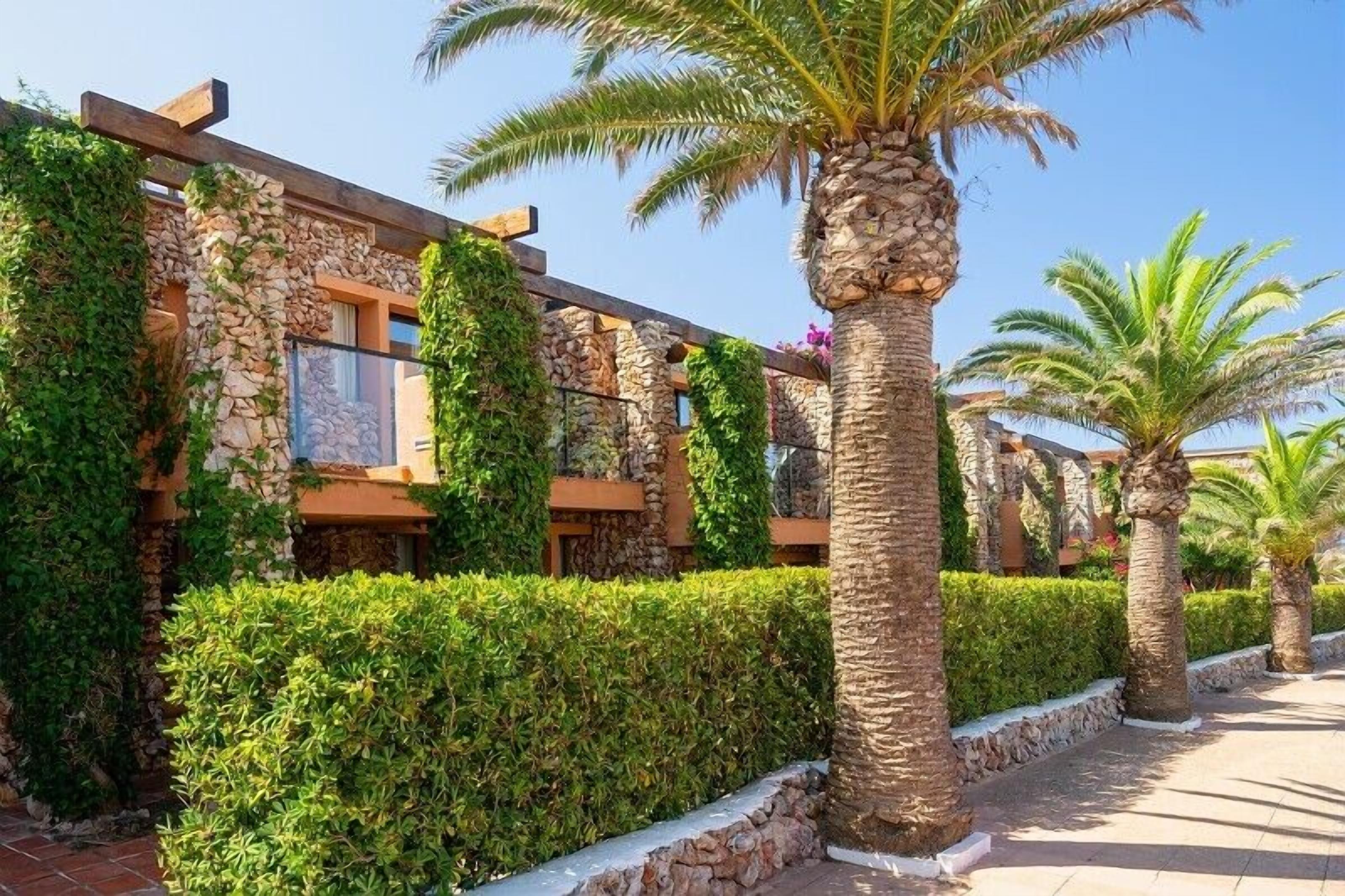 A building with stone walls and wooden beams, surrounded by well-maintained hedges and palm trees.