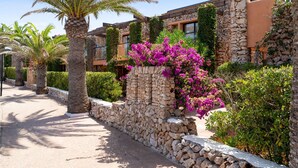 A stone pathway flanked by palm trees and vibrant bougainvillea flowers, leading to a building with stone walls and a modern design. - Hotel Menorca Binibeca  - Adults Recommended- by Pierre & Vacances Premium (Sant Lluis)