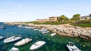 A coastal area with boats docked near rocky shores, a building with a balcony overlooking the water, and greenery on the hillside. - Hotel Menorca Binibeca  - Adults Recommended- by Pierre & Vacances Premium (Sant Lluis)