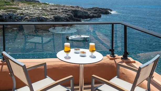 A glass-enclosed outdoor dining area with a table set for two, overlooking a rocky coastline and the sea. - Hotel Menorca Binibeca  - Adults Recommended- by Pierre & Vacances Premium (Sant Lluis)