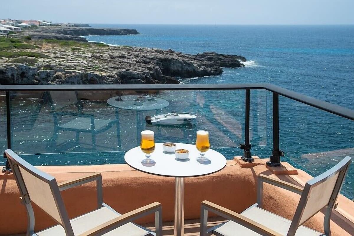 Un espace de restauration extérieur, abrité par des parois de verre, avec une table dressée pour deux personnes, offrant une vue sur une côte rocheuse et la mer.