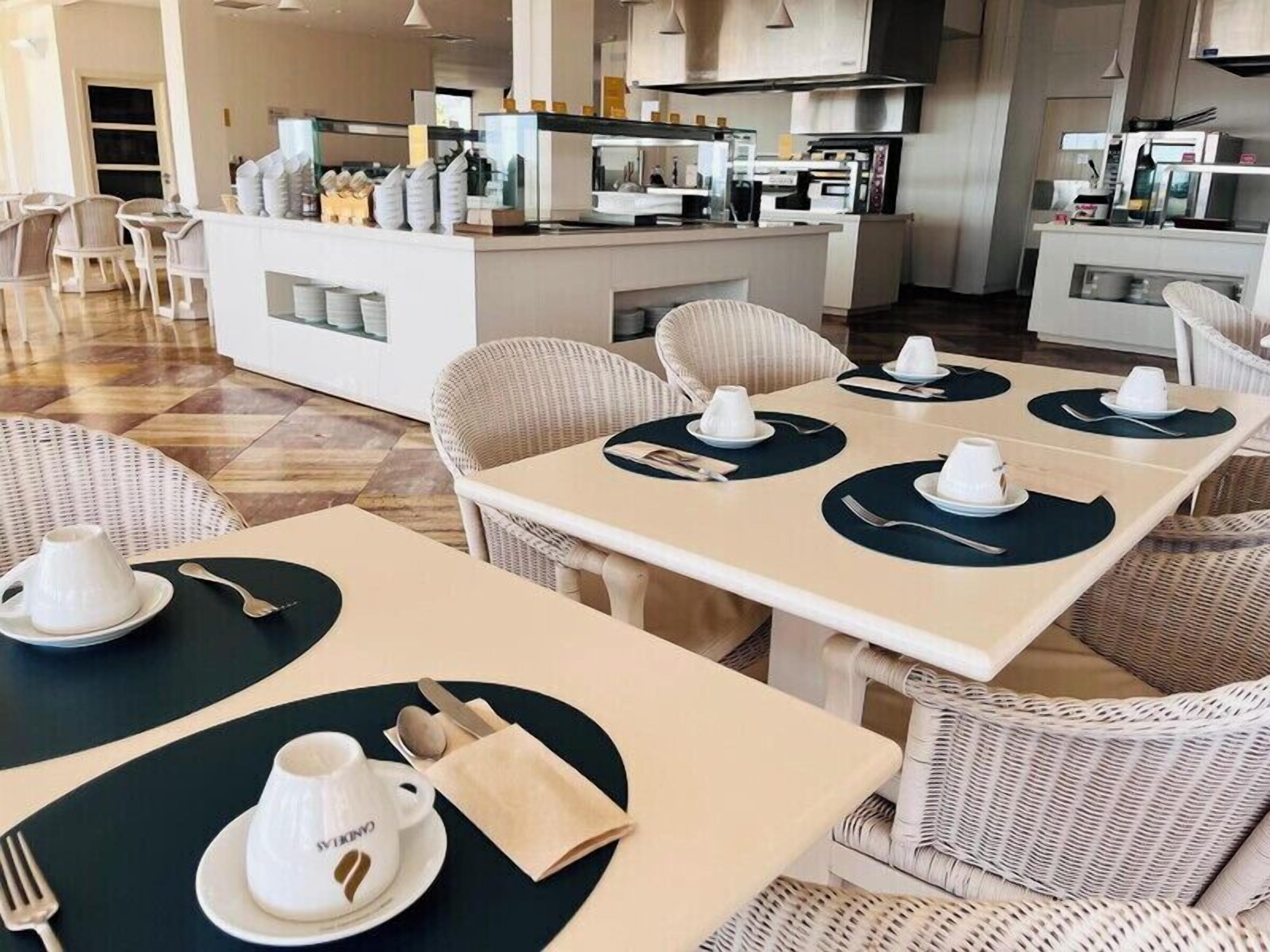 Een modern restaurant met witte rieten meubels, keurig gedekte tafels met witte kopjes en servetten, en een zichtbaar keukenhoekje op de achtergrond.