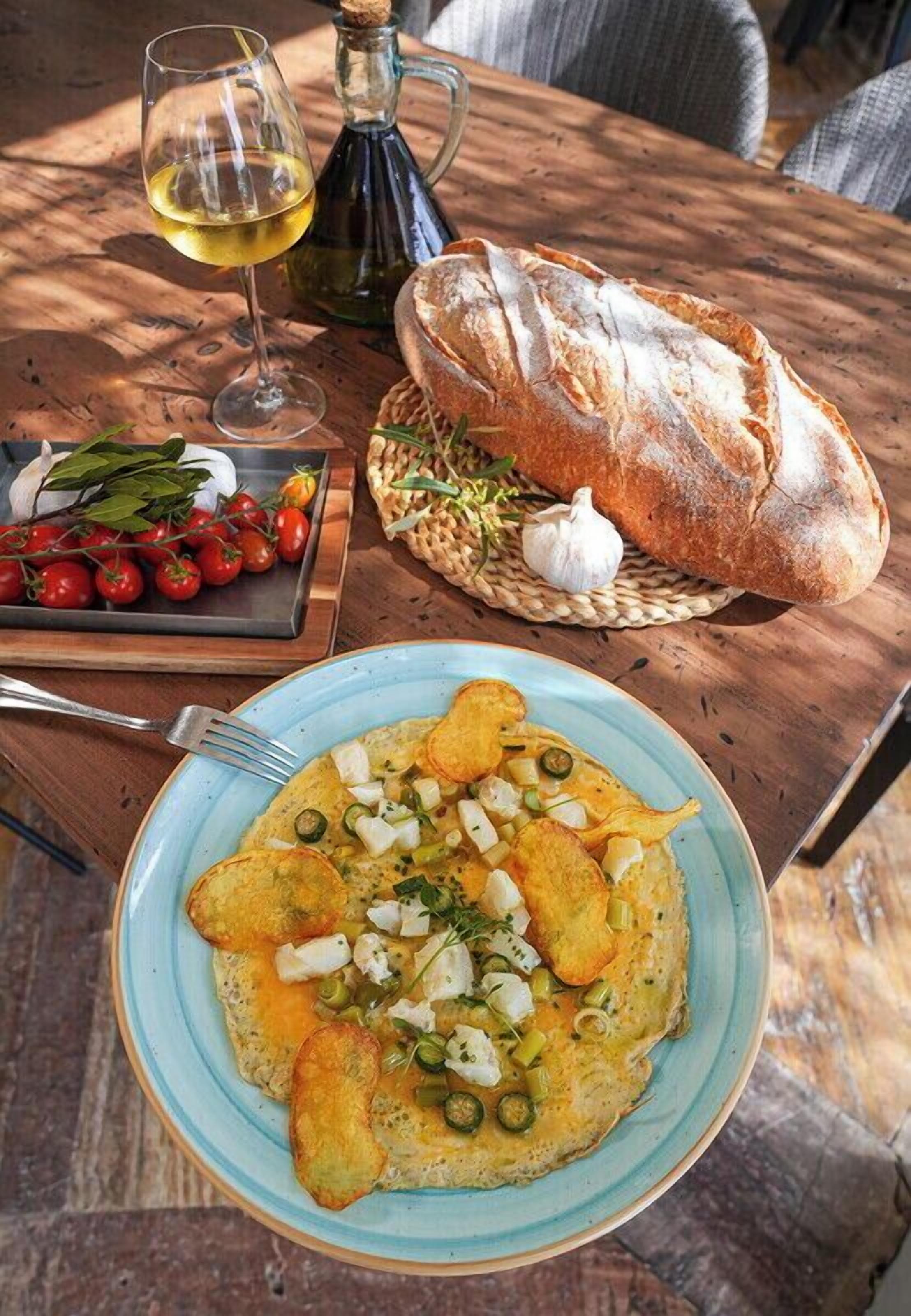 Una mesa con un plato de comida, una copa de vino, una botella de aceite de oliva, un pan y una cesta de tomates cherry y ajos.