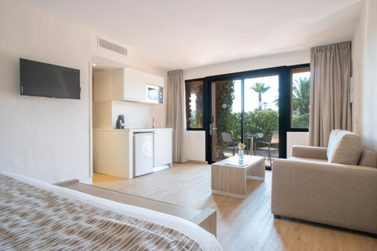 A modern hotel room with a bed, a sofa, a small kitchen area, and a balcony with a view of palm trees.