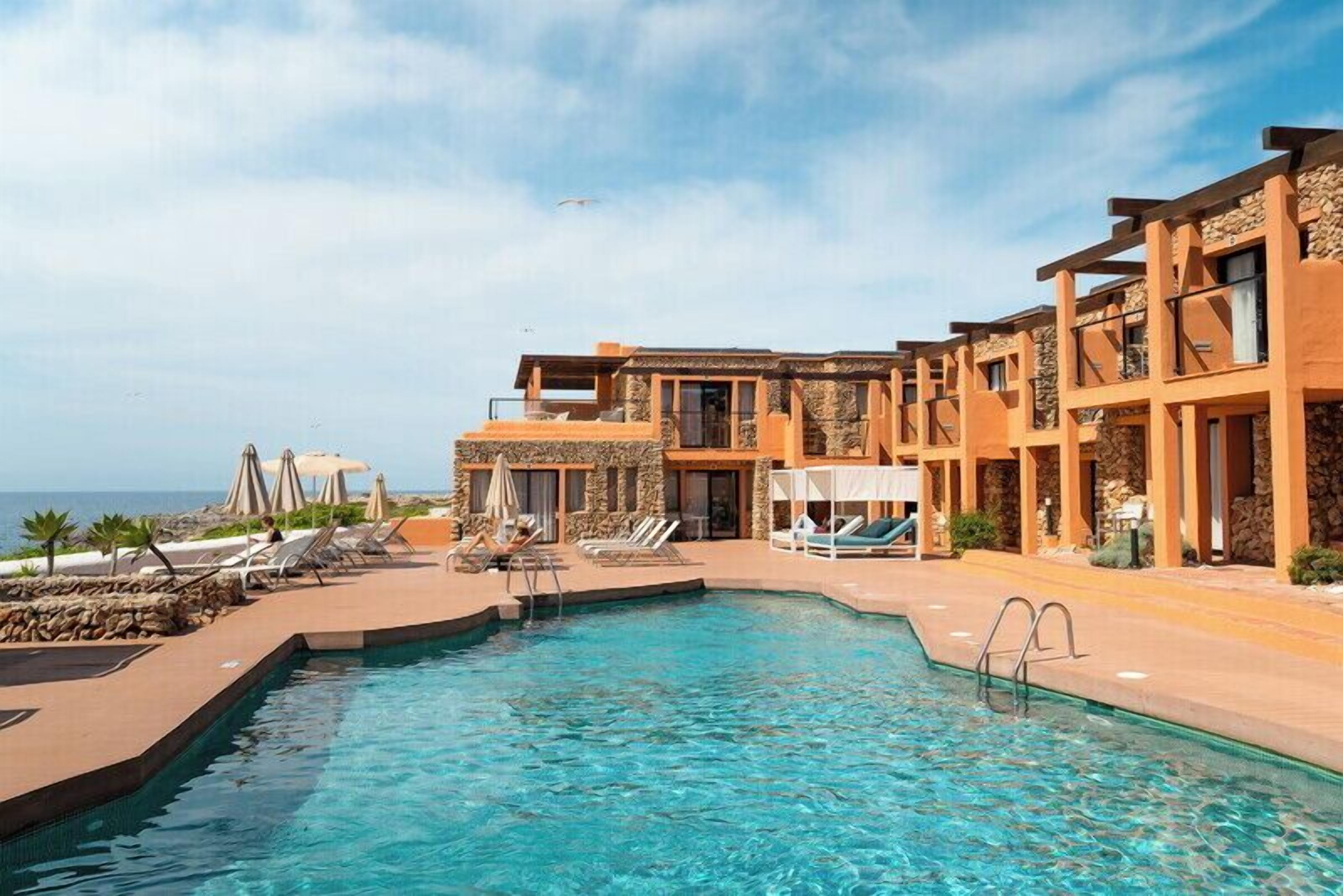 Un espace aménagé au bord d’une piscine à l’eau d’un bleu limpide, avec des chaises longues et un bâtiment dont la façade est agrémentée de pierres et qui possède des balcons.