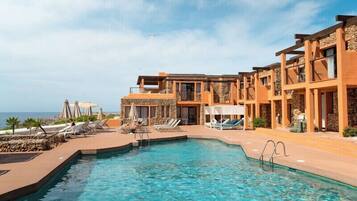 Un espace aménagé au bord d’une piscine à l’eau d’un bleu limpide, avec des chaises longues et un bâtiment dont la façade est agrémentée de pierres et qui possède des balcons.