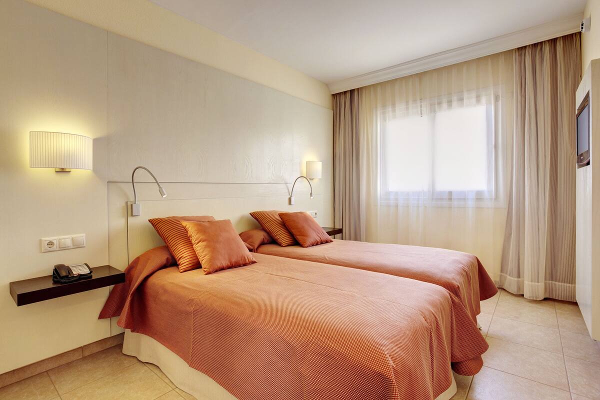 Une chambre d’hôtel avec deux lits, une table de chevet avec une lampe, un téléphone et une fenêtre avec des rideaux.