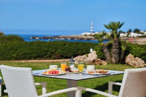 Une table dressée à l’extérieur avec des aliments pour le petit-déjeuner, une vue sur la mer et un phare à l’horizon.