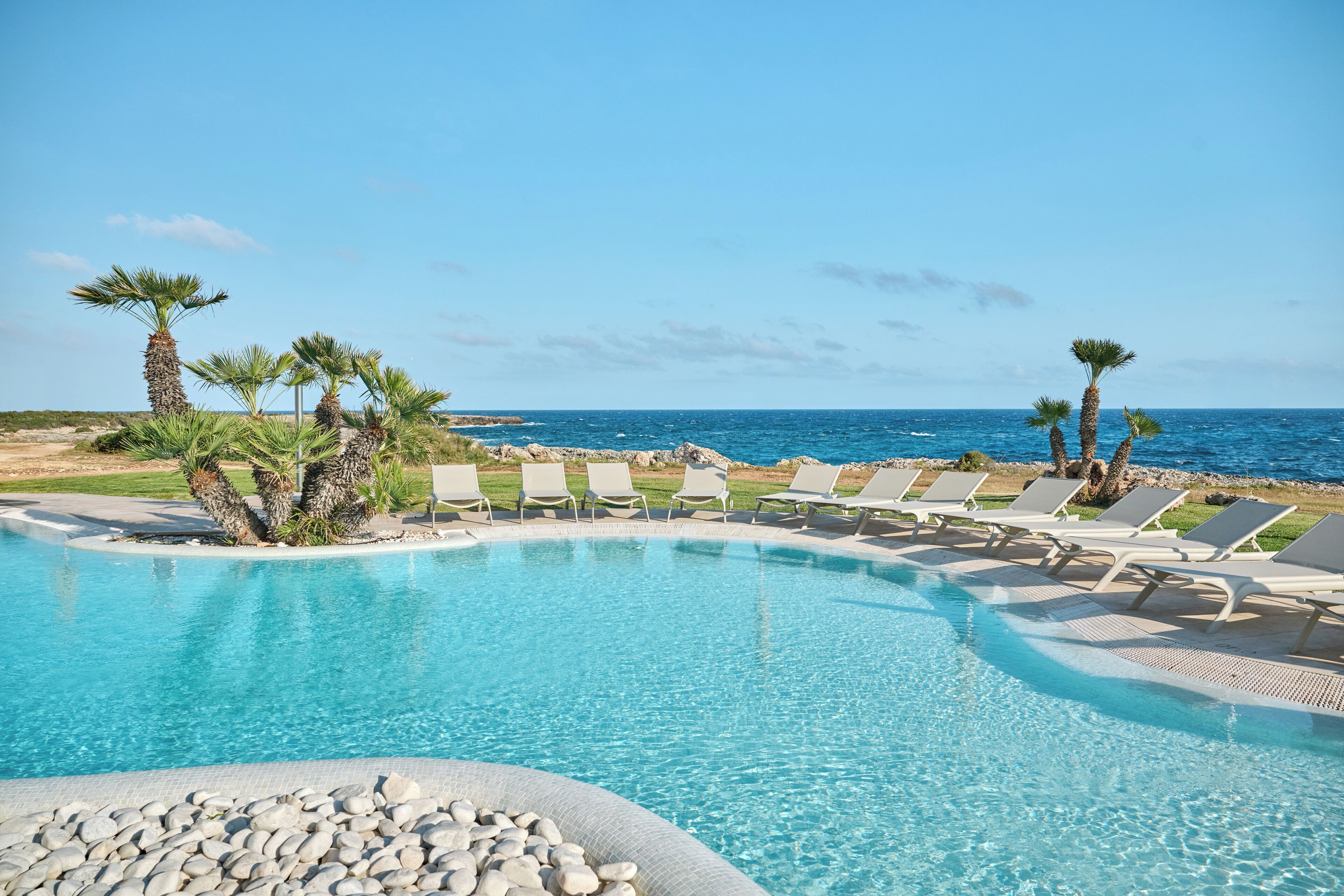 A clear blue pool with lounge chairs and palm trees, overlooking the ocean.