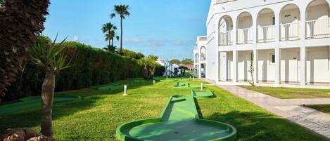 Un jardín cuidado con un putting de golf, palmeras y un edificio blanco con balcones arqueados.