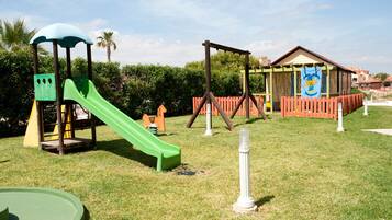 Un patio de juegos con un tobogán verde y columpios, rodeado por una zona vallada y un edificio al fondo.