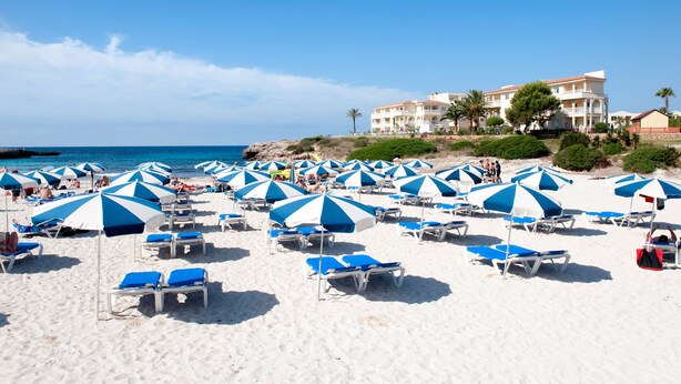 A beach with numerous blue and white umbrellas and lounge chairs, a building with multiple floors and balconies, and a clear blue sky. - Grupotel Tamariscos (Ciutadella de Menorca)