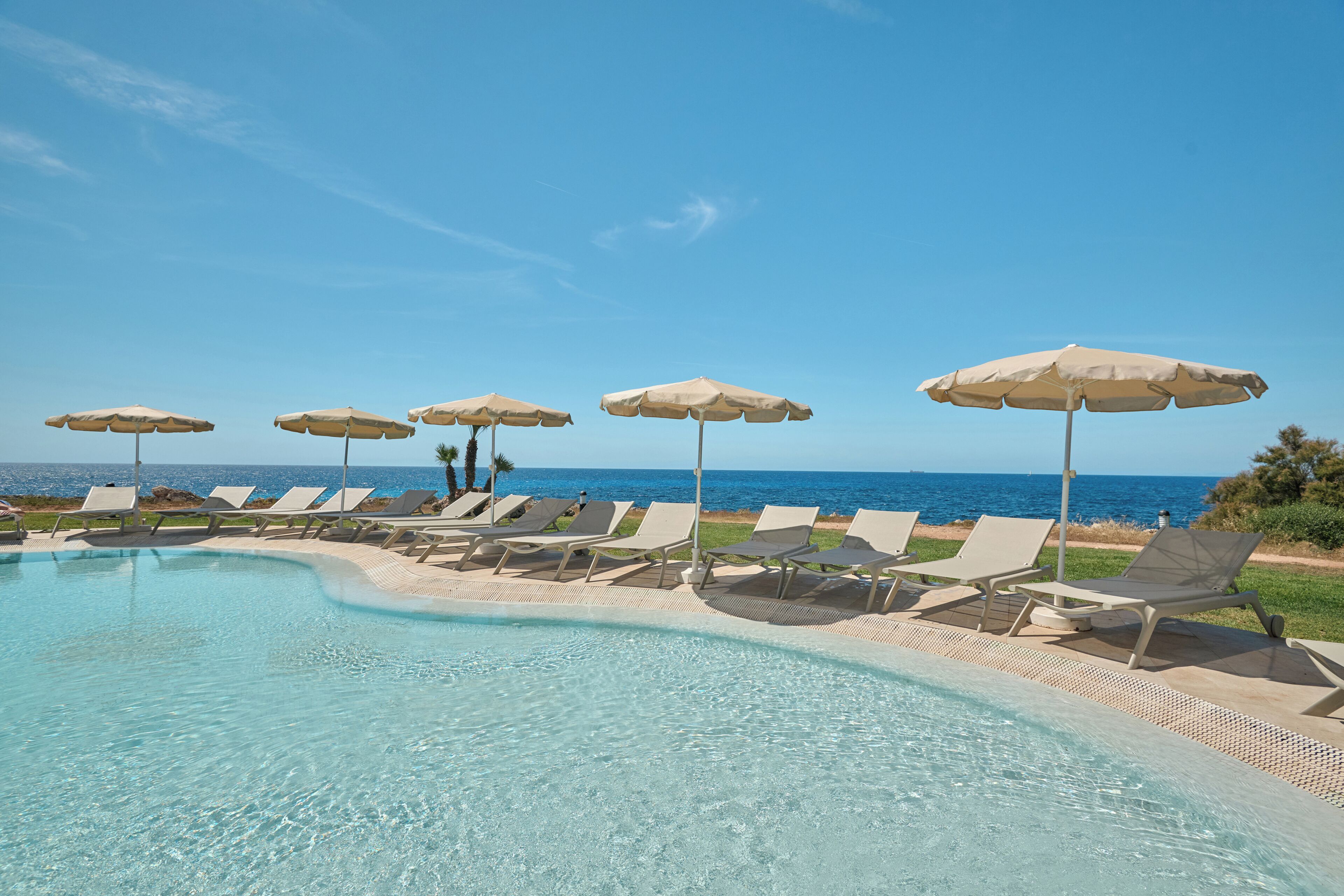 Zona junto a la piscina con sillones de descanso y sombrillas, con vista a un mar azul cristalino.