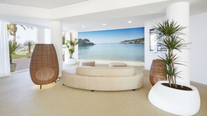 A modern living room with a large window overlooking a coastal view, a round sofa, and two wicker chairs. - Grupotel Tamariscos (Ciutadella de Menorca)