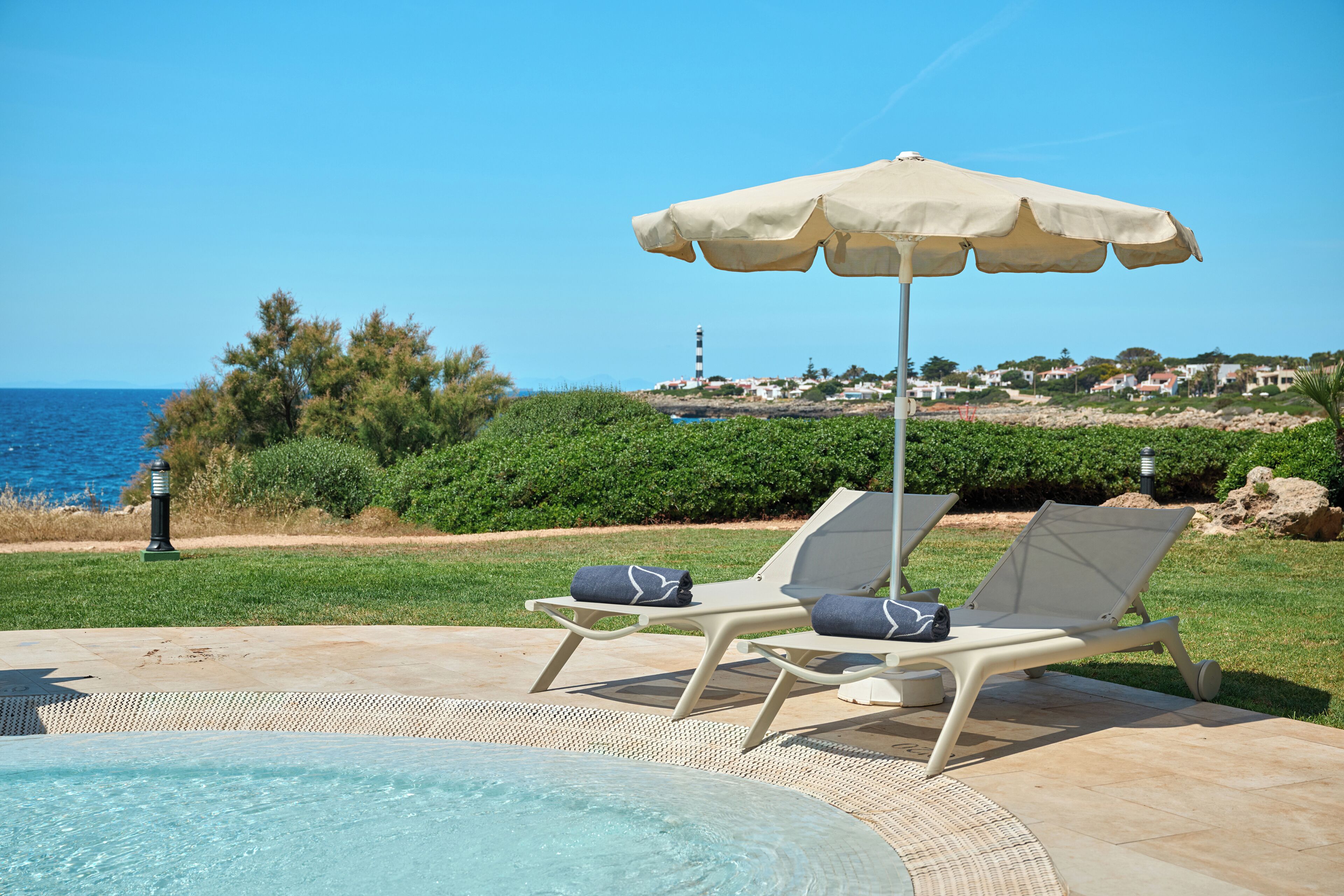 A pool area with two lounge chairs and an umbrella, overlooking a coastal landscape with a clear sky.