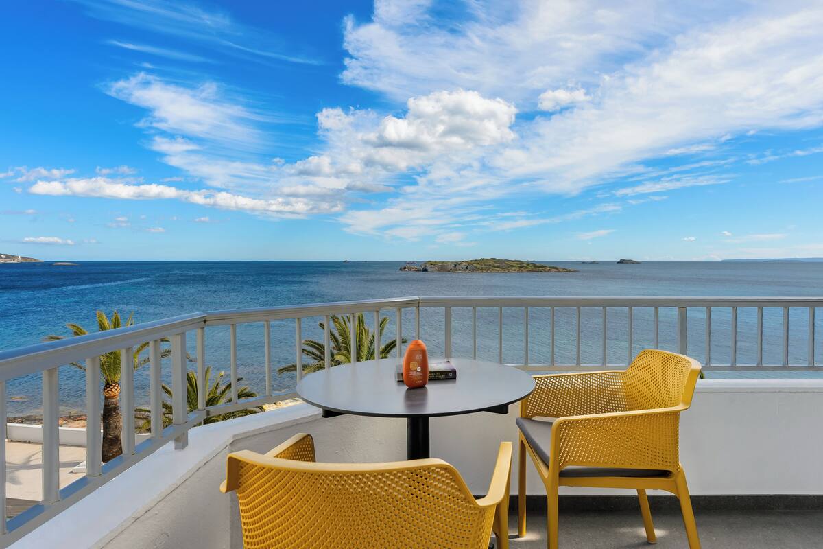 A balcony with a round table and two yellow chairs, overlooking a calm sea and a clear sky.