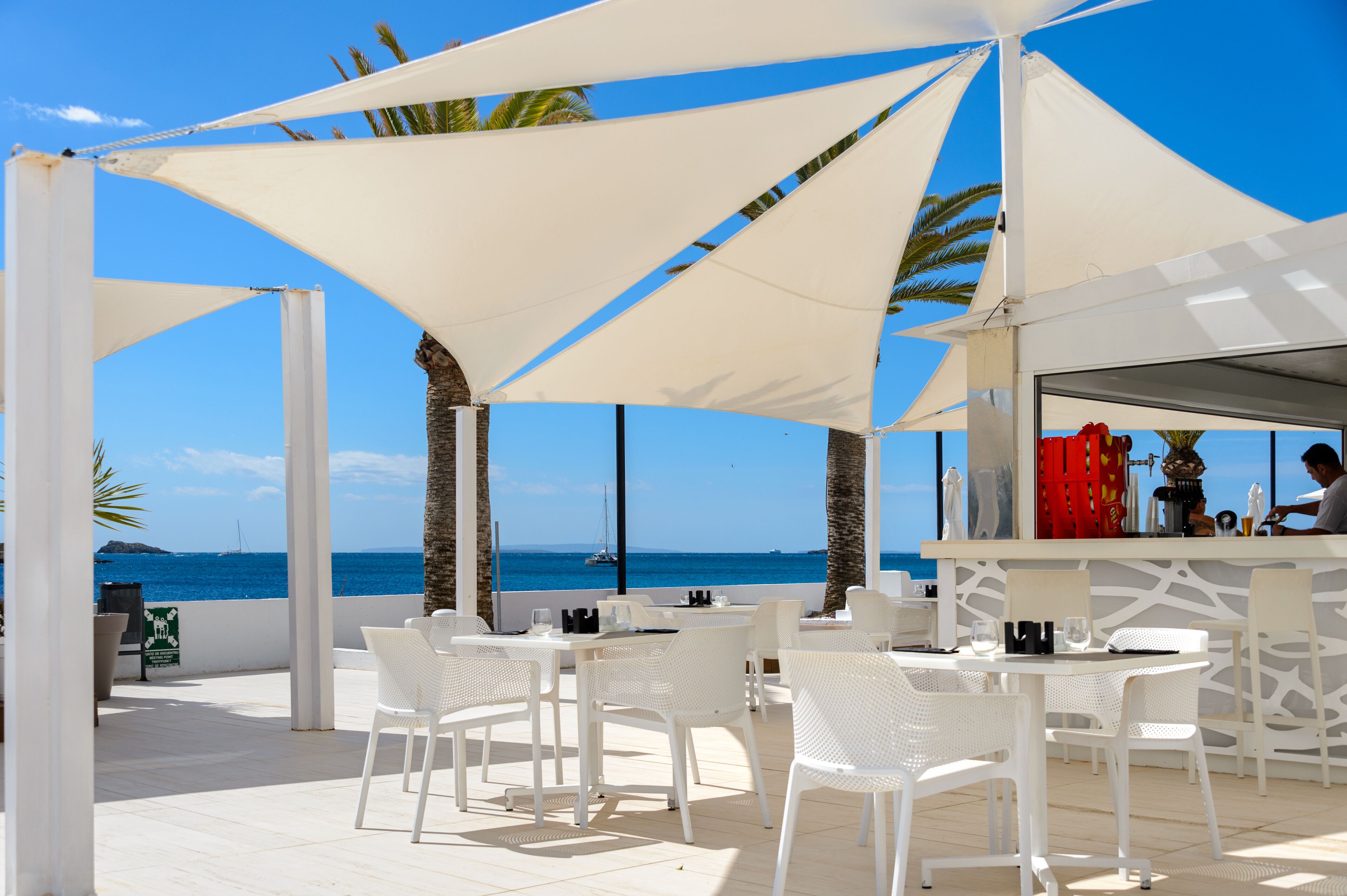 Un espace extérieur aménagé avec des chaises et des tables blanches, un bar et une vue sur la mer.