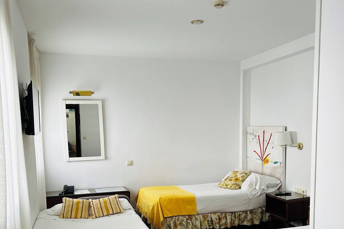 A hotel room with two single beds, a desk, a chair, a mirror, a lamp, and a ceiling light.