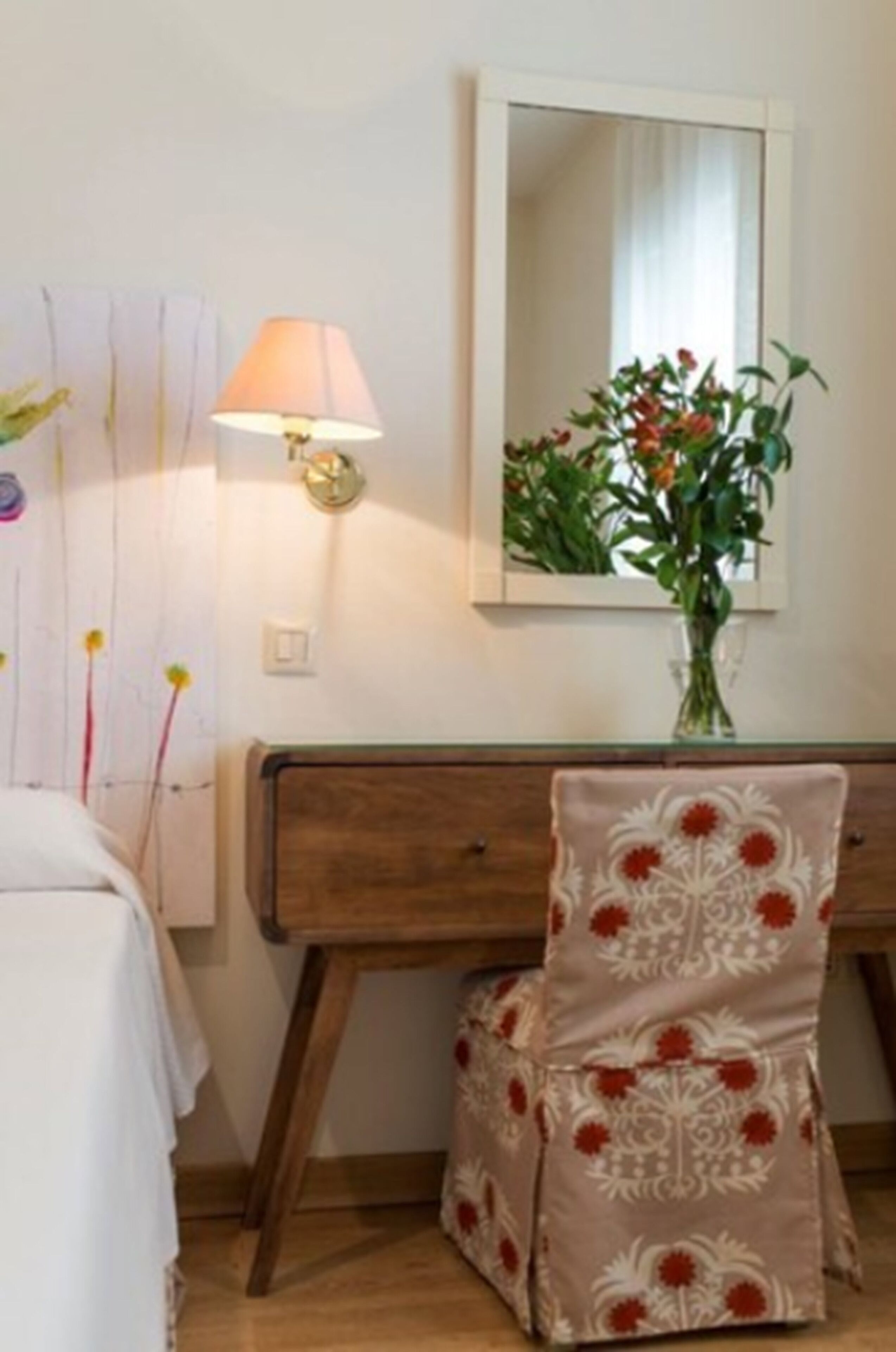 A bedroom with a bed, a bedside table, a lamp, a mirror, and a vase with flowers.