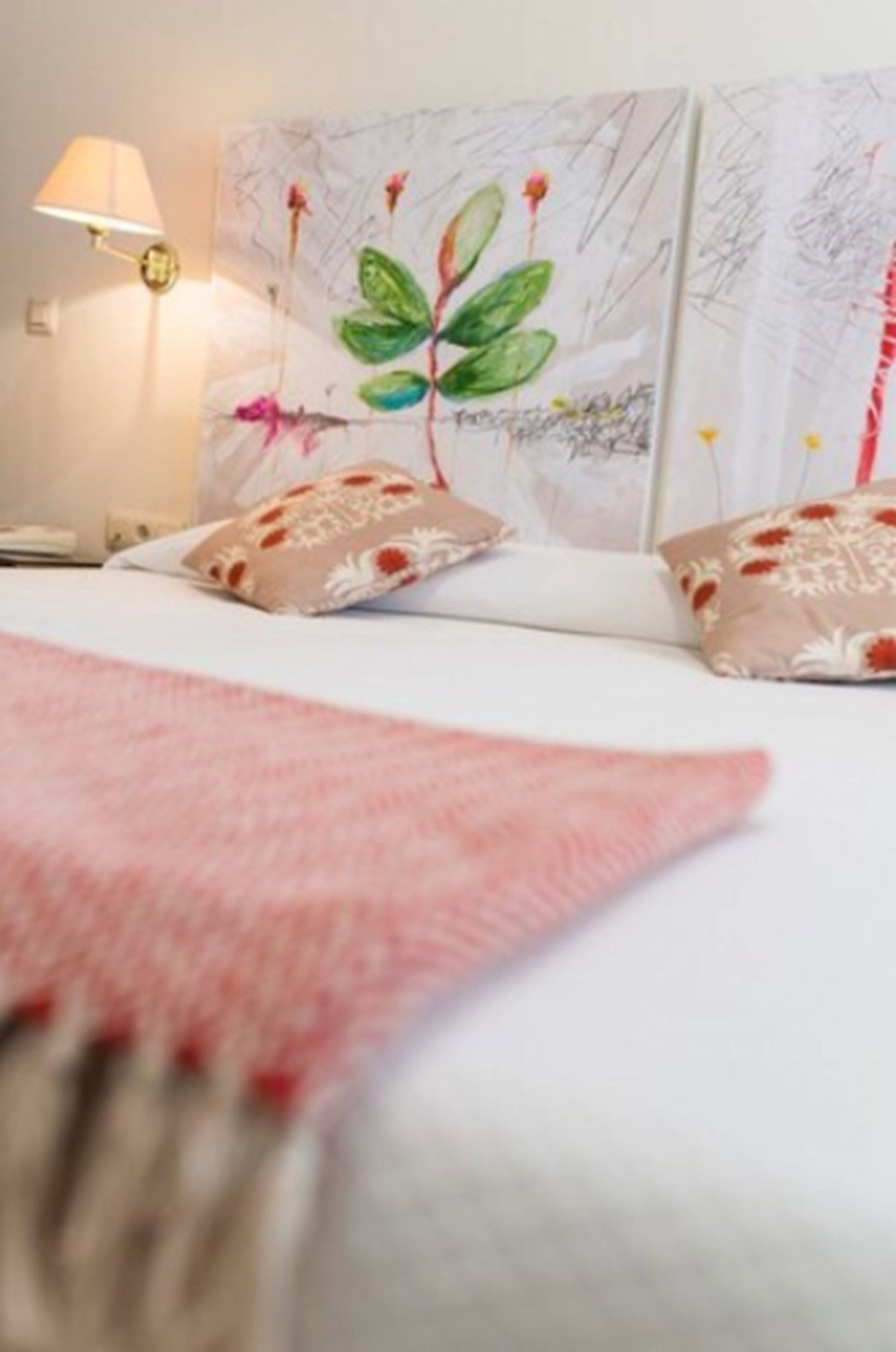 Uma cama bem arrumada com um cobertor rosa, parede com um quadro floral e uma luminária de cabeceira.
