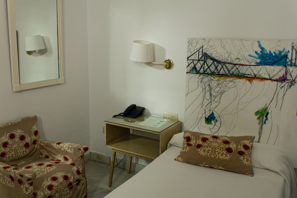 A hotel room with a bed, a desk with a lamp, a chair, and a wall with a painting.