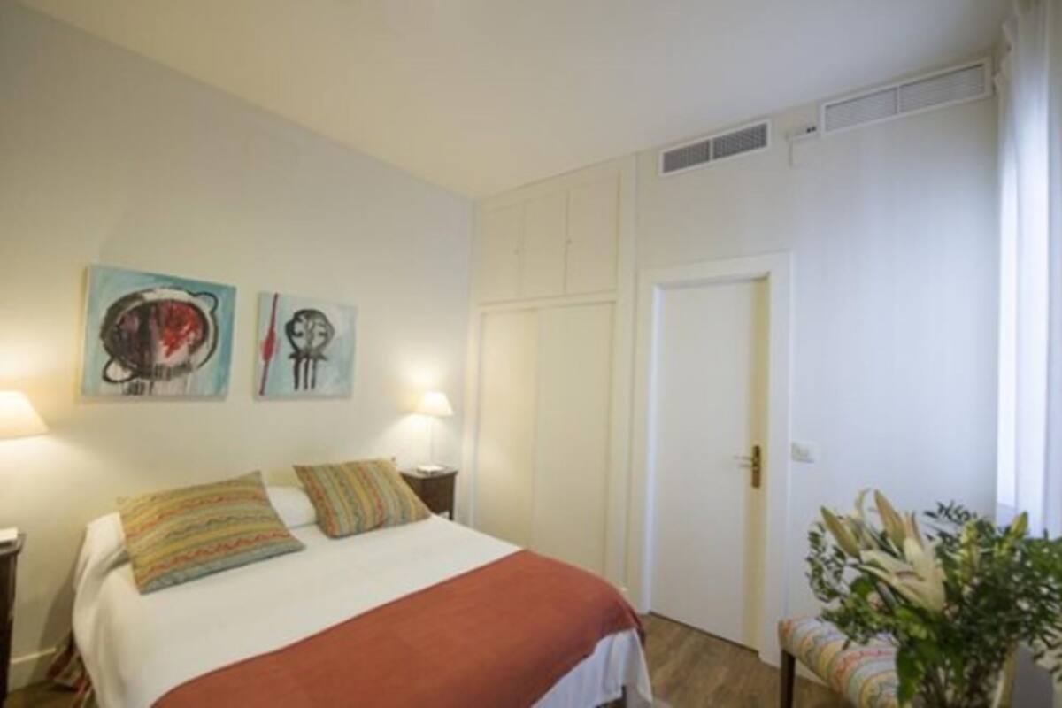 A bedroom with a bed, two wall-mounted lamps, and two paintings.