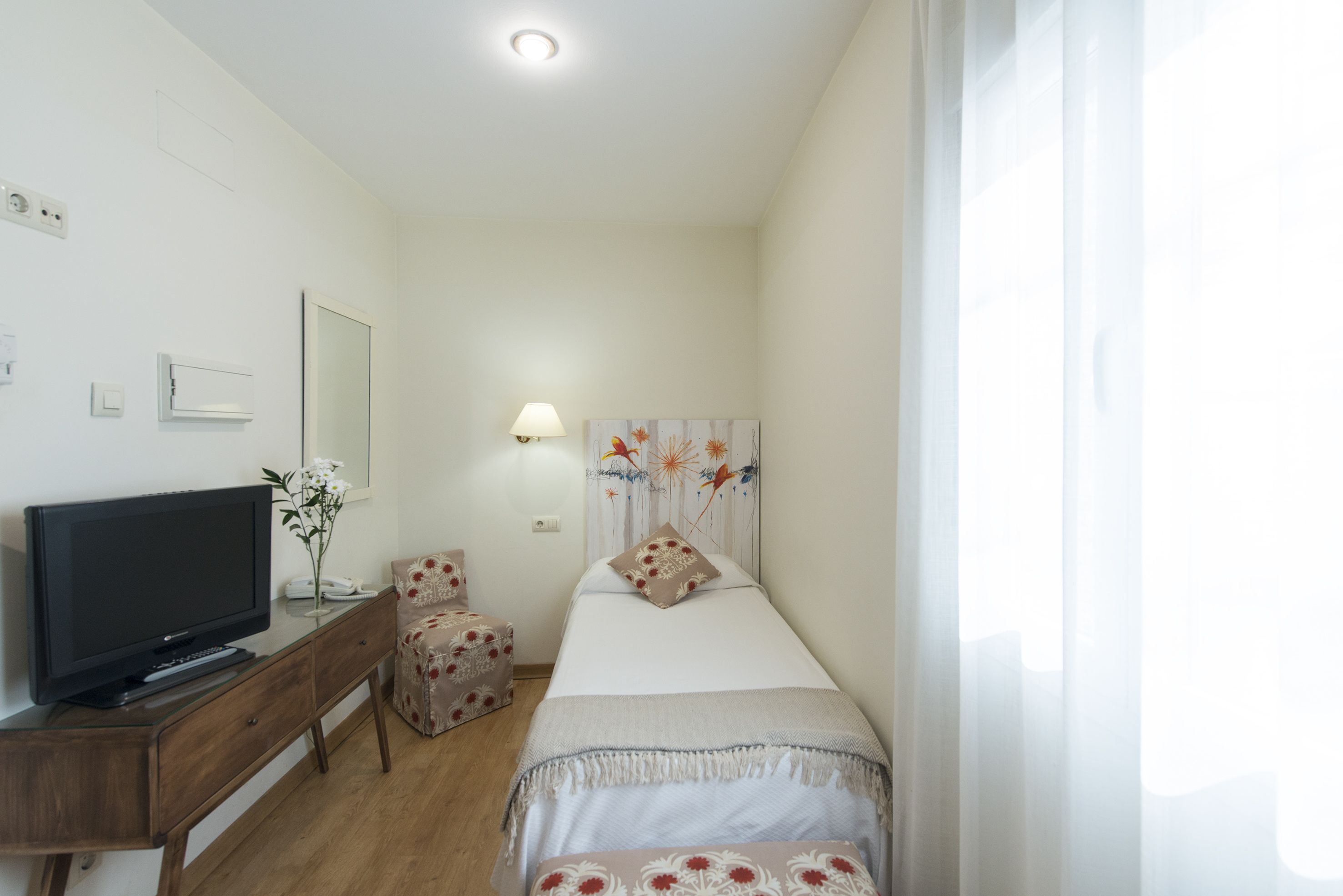 Un dormitorio con una cama, un televisor sobre un aparador de madera, una silla y una lámpara fijada a la pared.
