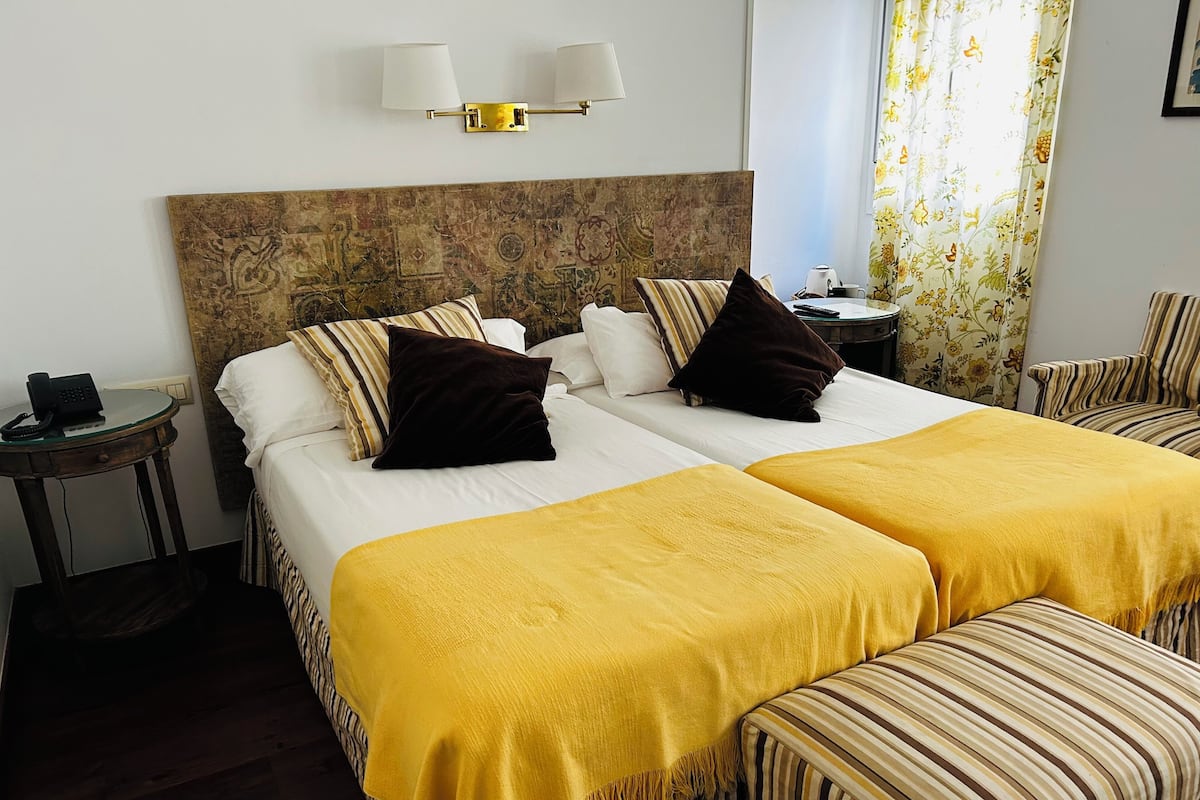 A hotel room with two beds, a headboard with a decorative panel, a bedside table with a phone, and a window with floral curtains.