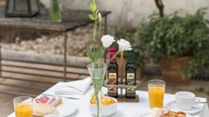 Mesa para desayuno al aire libre con mantel blanco, platos de comida, vaso de zumo de naranja, vaso de agua, botella de aceite de oliva y una vela.